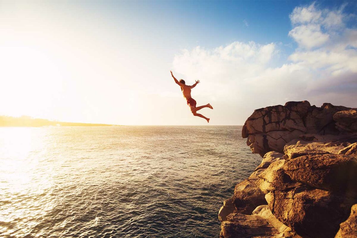 Man jumps off a cliff into the ocean at sunset.