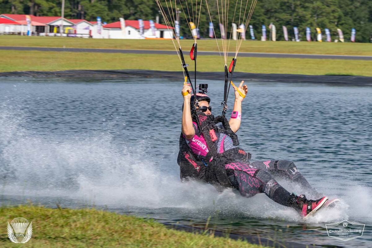 What is swooping in skydiving? Female skydiving swooper drags feet over the pond in a black jumpsuit with hot pink embellishment. 