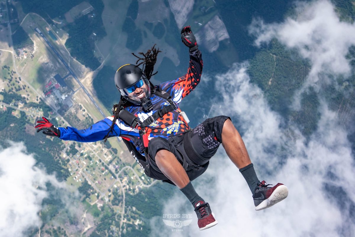 Male skydiver with dreadlocks in a sit position and clouds in the background. 