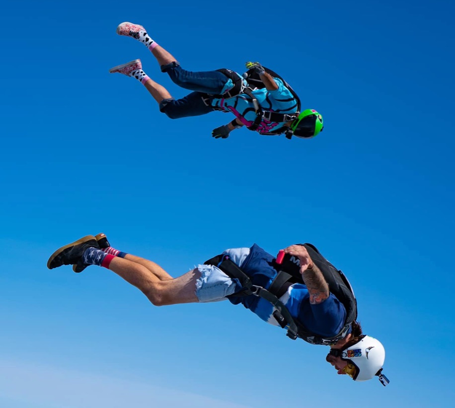 Two skydivers angle flying in front of the bright blue sky.